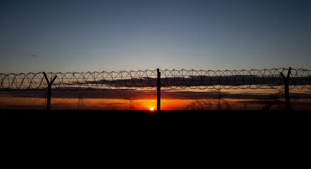 Obraz premium Silhouettes of birds flying over a barbed wire fence at sunset