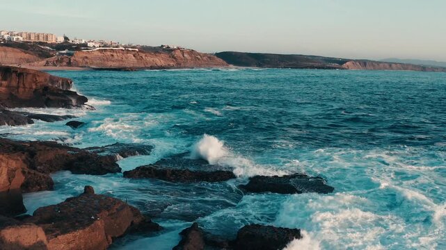 Aerial view: Large ocean waves crash against the rocky shore during sunset. Drone: Scenic view of the sea coast in summer at sunset. Beautiful seascape with crashing waves on rocks. Ericeira, Portugal