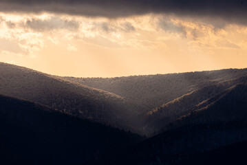 Obraz premium Atmospheric winter mountain slopes covered with snow and frost at sunset, golden light rays illuminating foggy forest valleys, moody mountain landscape with soft sunlight breaking through clouds.