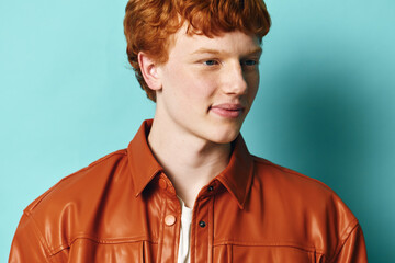 Young man with red hair wearing a brown leather jacket standing against a blue background. Portrait...