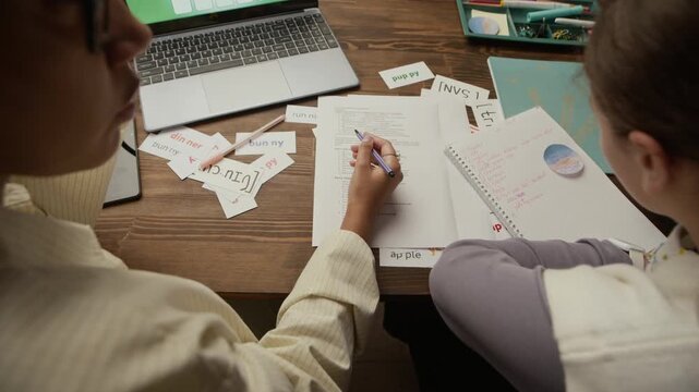 High angle shot of unrecognizable tutor and teen student sitting at table, woman explaining theorem while teenager writing it in notebook