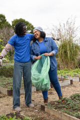 African American couple tending wooden raised beds on gravel path, holding translucent green bag