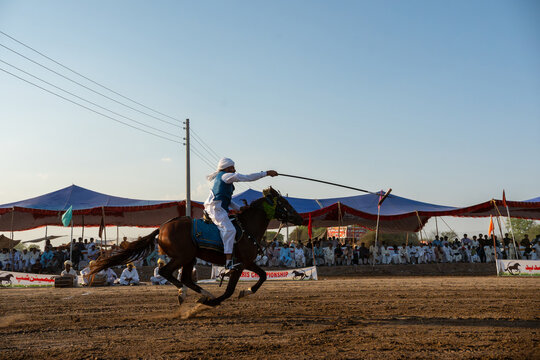 Punjab, Pakistan - 24 April 2024: View of a skilled horseman, adorned in traditional attire, executes a daring stunt with a spear amidst a lively crowd.