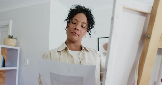 Adult African American woman holding sheet, matching canvas on easel in home studio, evaluating fit
