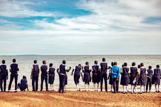 Kampala, Uganda - 26 March 2024: View of students lined up on the sandy beach, gazing out at the serene water meeting the horizon under a partly cloudy sky.