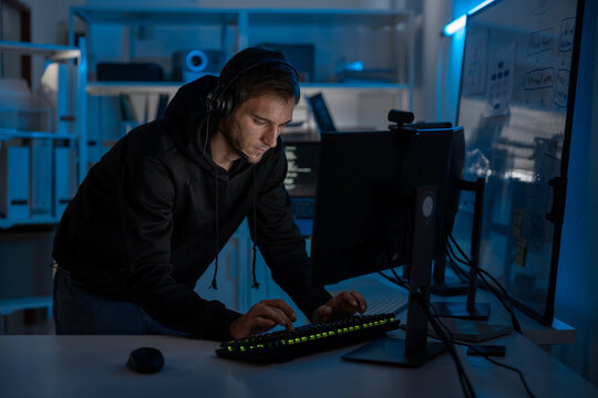 Male programmer and developer focused on computer screen at night, coding digital systems and ensuring cyber security. Freelancer with headset working overtime under blue light in office.