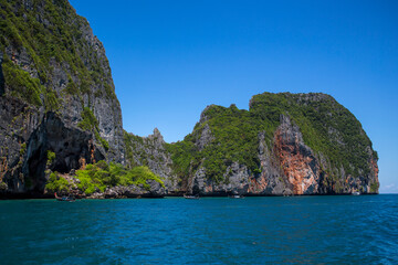 Fototapeta premium Tropical bay enclosed by massive limestone cliffs. The steep rock faces are partially covered in dense green foliage and show various shades of grey and orange. The deep blue sea in the foreground.