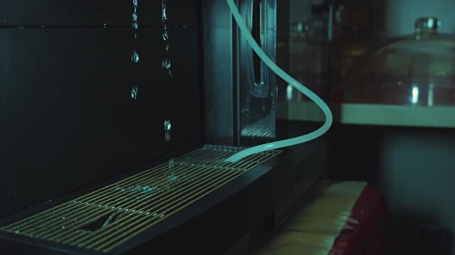 Close-up of a modern espresso machine's cleaning cycle. Water drips onto the metal tray in a dark, moody kitchen. The cool, low-key lighting highlights the droplets and sleek machine details