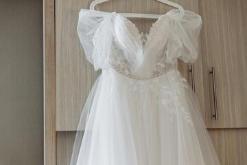 White lace wedding dress with tulle sleeves hanging on a wardrobe, beautiful bridal gown detail before the ceremony