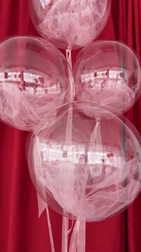 Four transparent bobo balloons filled with soft pink tulle fabric, hanging against a rich red velvet curtain. Elegant and trendy festive decoration