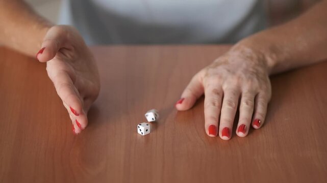 Close-up of female hands with vitiligo and red manicure placing dice on a wooden table. Concept of chance, risk, gambling, uncertainty, and inclusive beauty. Indoor lifestyle scene with focus on hands