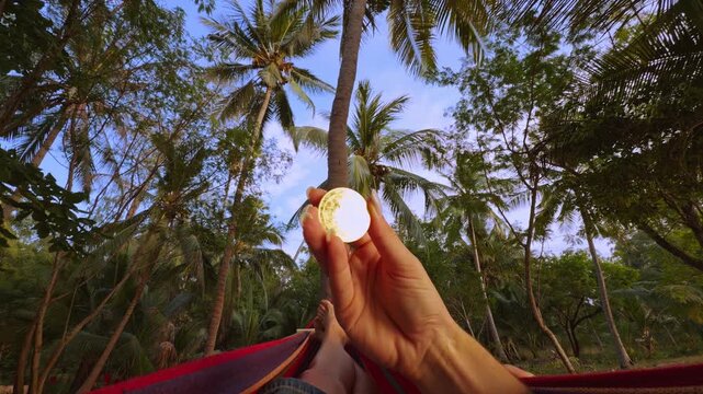 First Person View Holding Golden Bitcoin Coin While Resting in Hammock Among Palm Trees in Tropical Forest Showing Digital Wealth and Relaxed Travel Lifestyle