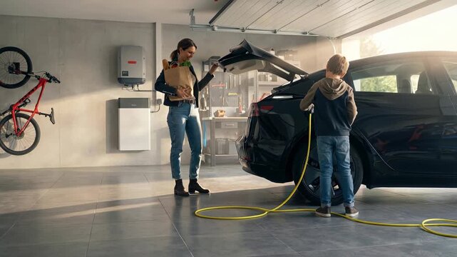 Wide shot of a young boy plugging a yellow charging cable into a black electric car in a modern home garage while his mother stands nearby holding a grocery bag of fresh produce.