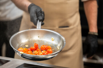 A professional chef in black gloves and a tan apron tosses cherry tomatoes and garlic in a stainless steel frying pan while cooking in a commercial kitchen