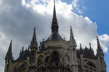 Fototapeta premium Ornate stone spires and intricate flying buttresses reach toward the dramatic clouds above the historic Reims Cathedral. Carved arches and statues frame darkened windows.