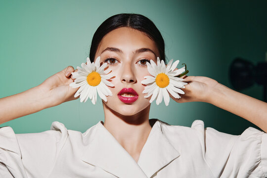 Young woman with dark hair holds two large white daisies in front of her face wearing bright pink lipstick and white top. Glossy makeup highlights the creative beauty fashion concept.