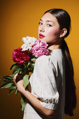 Young woman with glossy dark hair and red lipstick holds bright peony flowers against yellow...