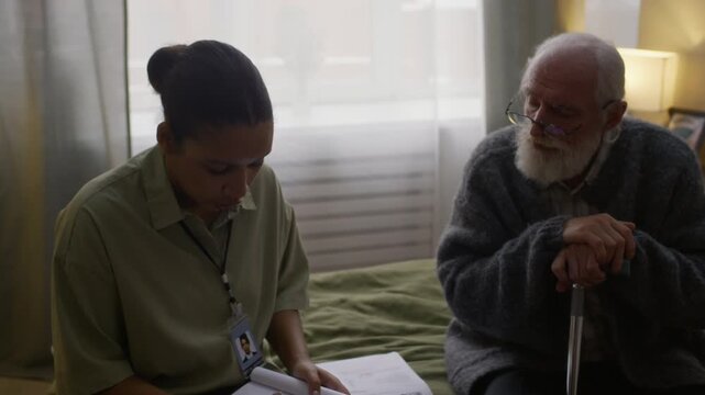 Handheld zoom-out shot of young Black female social worker filling in questionnaire form, assessing needs and living conditions of deprived senior Caucasian man sitting on bed at home