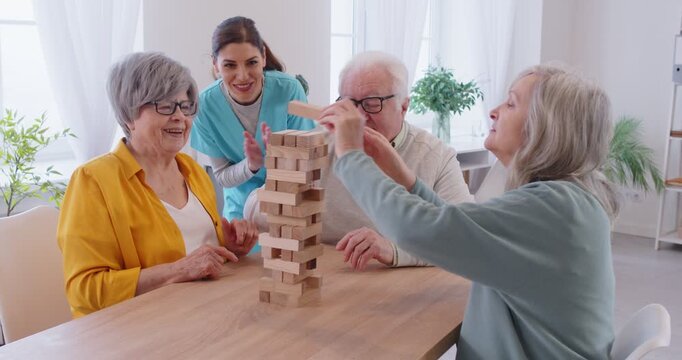 Group of elderly people sitting and playing board game together, taking blocks from wooden tower at table. Joyful seniors playing jenga during leisure activity with young female nurse cheering them.