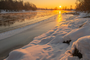 Golden rays of the sun break through the chilly morning air, reflecting on the icy surface of the river. Freshly fallen snow blankets the ground, creating a serene winter landscape