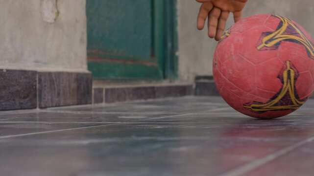 Black child hand rolling red ball across tiled floor, closeup of scuffed rubber and faded logo, doorway and courtyard backdrop, dynamic momentum and soft bounce, casual neighborhood play