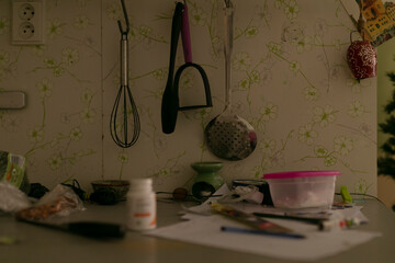 Cluttered counter with bowl and pill bottles, hanging utensils and scattered papers, dim lighting casting soft shadows, worn wallpaper background, intimate tired atmosphere © Viktar