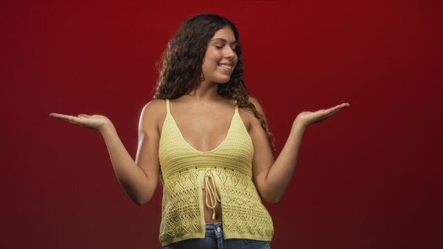 Woman in a yellow crochet top and jeans with exposed midriff, palms up presenting and smiling in a red studio; playful confidence.