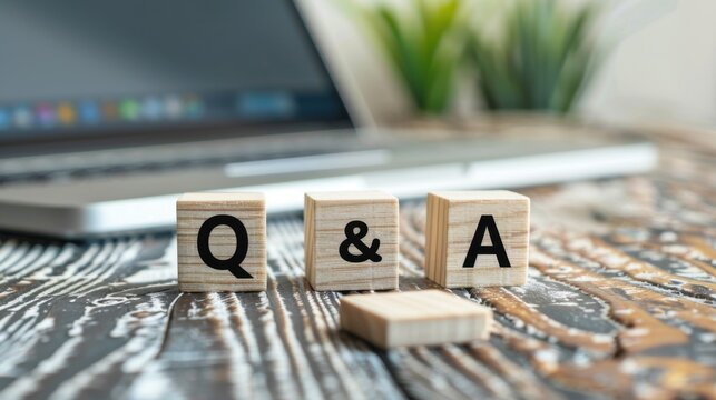 Wooden blocks with letters Q and A on a rustic table. A laptop and green plants are in the background, suggesting a workspace or learning environment.