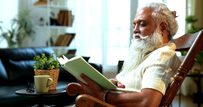 Indian mid age bearded man reading book novel on relaxing chair at home, stylish mature male enjoying peaceful leisure time with printed copy in modern interior lifestyle setting