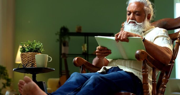 Indian mid age bearded man reading book novel on relaxing chair at home, stylish mature male enjoying peaceful leisure time with printed copy in modern interior lifestyle setting