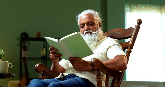 Indian mid age bearded man reading book novel on relaxing chair at home, stylish mature male enjoying peaceful leisure time with printed copy in modern interior lifestyle setting