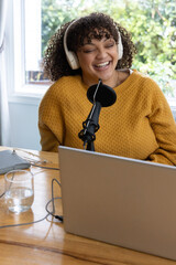 African American woman wearing mustard sweater, recording podcast at wooden table with microphone