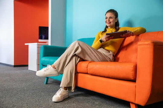 Woman sitting on orange sofa holding tablet, wearing mustard top, smartwatch and glancing in office