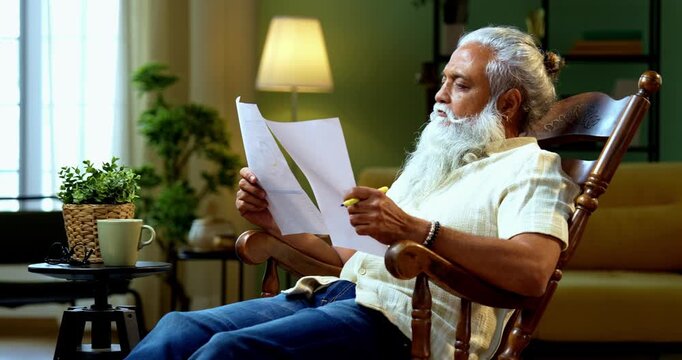 Indian mid age bearded man reading and showing financial document to camera sitting on relaxing chair at home, stylish mature male evaluating profit and loss report with positive expression