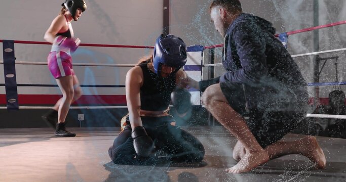 Kneeling boxer wearing blue helmet and black gloves pausing in gym ring, coach barefoot tending