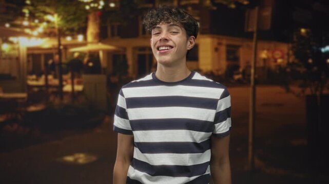 Teenager boy in striped shirt makes ok sign with hand over eye and smiles while standing on a night street outside a cafe; playful mischief.