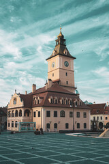 Fototapeta premium The historic Council House in Brasov stands proudly against a bright, streaked sky. Its iconic clock tower overlooks a wide, patterned cobblestone square, capturing the timeless soul of Transylvania.