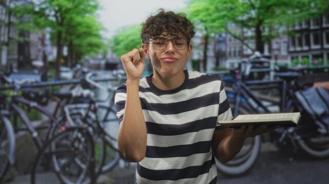 Man student holds hardcover book with one hand and touches temple with finger while smirking in street; study focus contemplation.