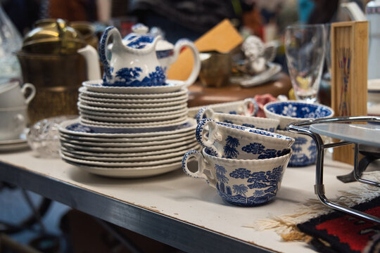Blau‑wei&szlig;es Porzellanservice mit Tellern, Tassen und Kannen auf Marktstand. Hand greift nach kleinem Krug. Detailreiches Vintage‑Geschirr in dichter Anordnung.