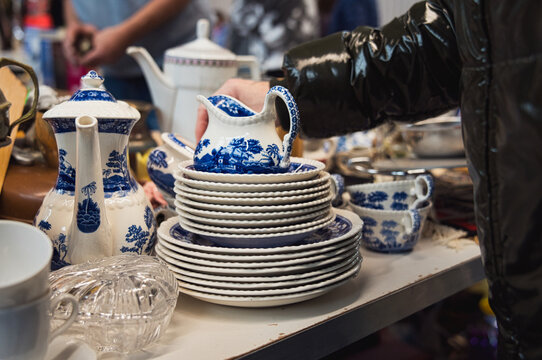 Blau‑wei&szlig;es Porzellanservice mit Tellern, Tassen und Kannen auf Marktstand. Hand greift nach kleinem Krug. Detailreiches Vintage‑Geschirr in dichter Anordnung.