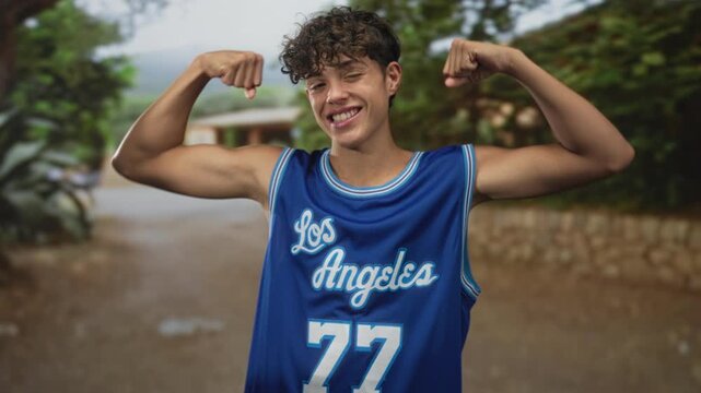 Teen boy flexes biceps and shows smile while wearing blue los angeles basketball jersey in street with stone wall and trees; youthful confidence.