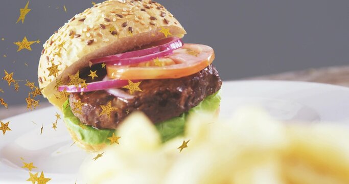 Sesame-seed hamburger sitting on white plate at tabletop, with gold star confetti and blurred fries