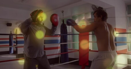 Training coach holding red focus mitts while shirtless boxer throwing punch in ring, window, gloves