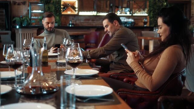 Group of friends sitting at restaurant table but ignoring each other, all using mobile phones during lunch. Concept of social isolation and smartphone addiction in modern society.