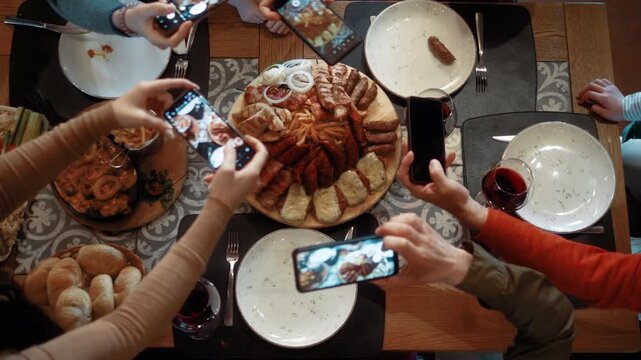 Top view of dining table with served food while multiple hands photograph dishes using smartphones. Overhead shot capturing modern food sharing and digital lifestyle in restaurant.