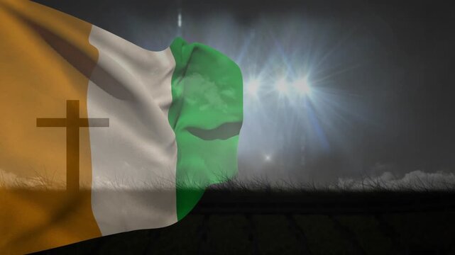 Wind rising, orange-white-green flag billowing and curling under floodlights, revealing faint cross