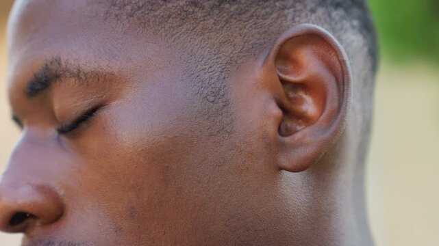 Black male athlete closeup face sweating, eyes closed during postsprint recovery, droplets on cheek glisten in sunlight, focused breath control, sprinter cooling down after interval, outdoor training