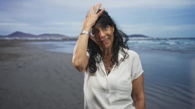 Woman with hand to head touching hair at beach wearing white shirt and silver bracelet near shoreline; serene reflection.