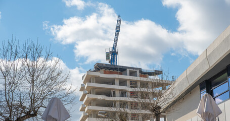 Modern concrete building under construction with a blue crane against a cloudy sky. Urban development, architecture, and civil engineering project in progress © John_Doo78