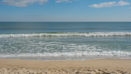 Obraz premium Wide shot of a serene sandy beach with gentle white waves rolling onto the shore under a clear blue sky. Peaceful coastal landscape with a distant horizon line